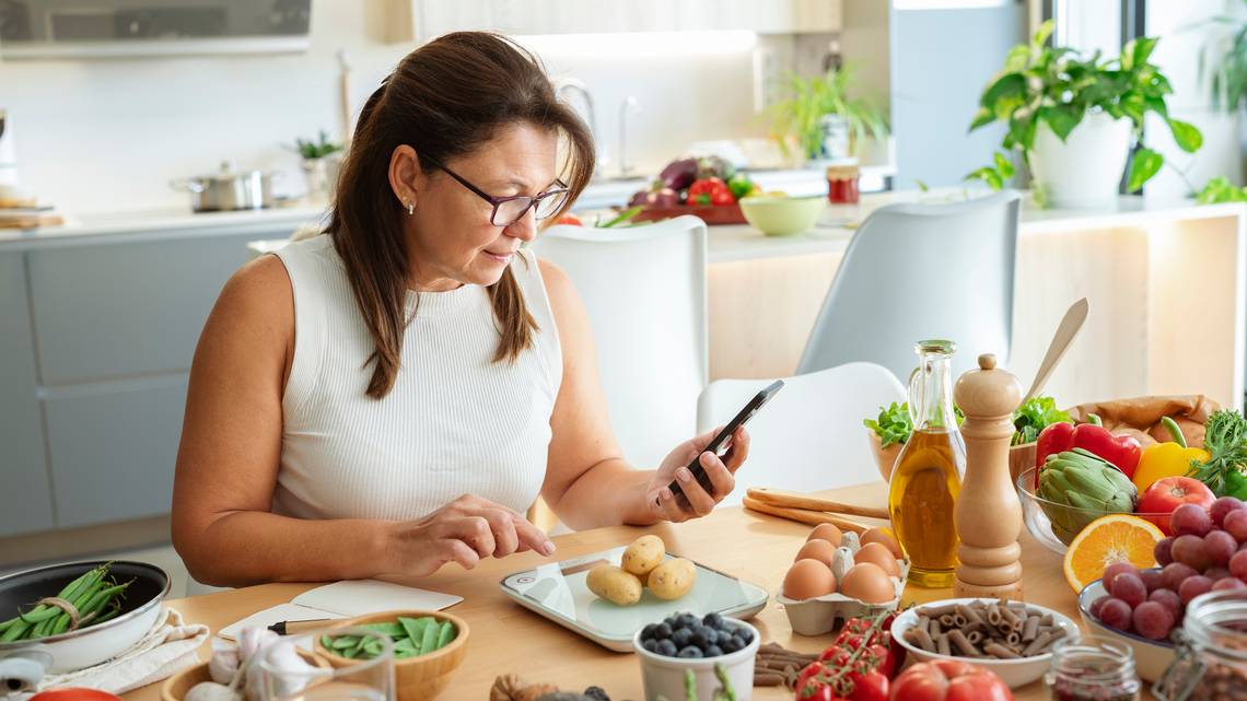 A woman sits in a bright kitchen using her smartphone while weighing potatoes on a food scale, surrounded by fresh fruits, vegetables, eggs, and pantry staples—illustrating mindful meal planning and nutrition tracking, ideal for a Cronometer user.
